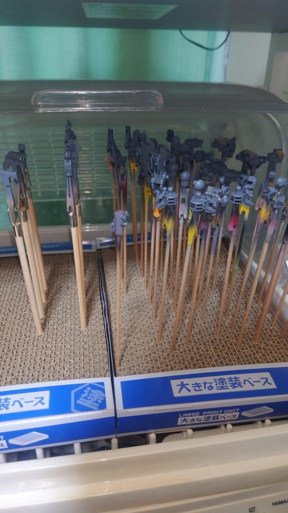 では実際に塗装。クセというか作業のしやすさから、大体はフレームから塗装しています。フレームからなのは滅多に塗分けせず1色で終わるから。30分もあれば上塗りできる状態です。一通り塗ったら30分は軽く過ぎているので、すぐにトップコートを吹いて終わり。ちなみにパーツの持ち手は・クレオス ネコの手持ち手棒 通常&ミニクリップ・ホビーベース 持ちやすい塗装棒 逆作用タイプ・プラモ向上委員会 塗装クリップMaster・竹串+両面テープをパーツの保持しやすい箇所&塗装に影響のない箇所で保持します。また個人的にフレーム系で重要なのは「関節部をマスキングテープで保護」することです。これをやらないと組み合わせたときに入りづらいのは序の口、最悪は塗膜のせいで動かない、無理に動かした結果可動部がバキ折れます。私の作品群では関節が動かない・動きにくいのは恒例です