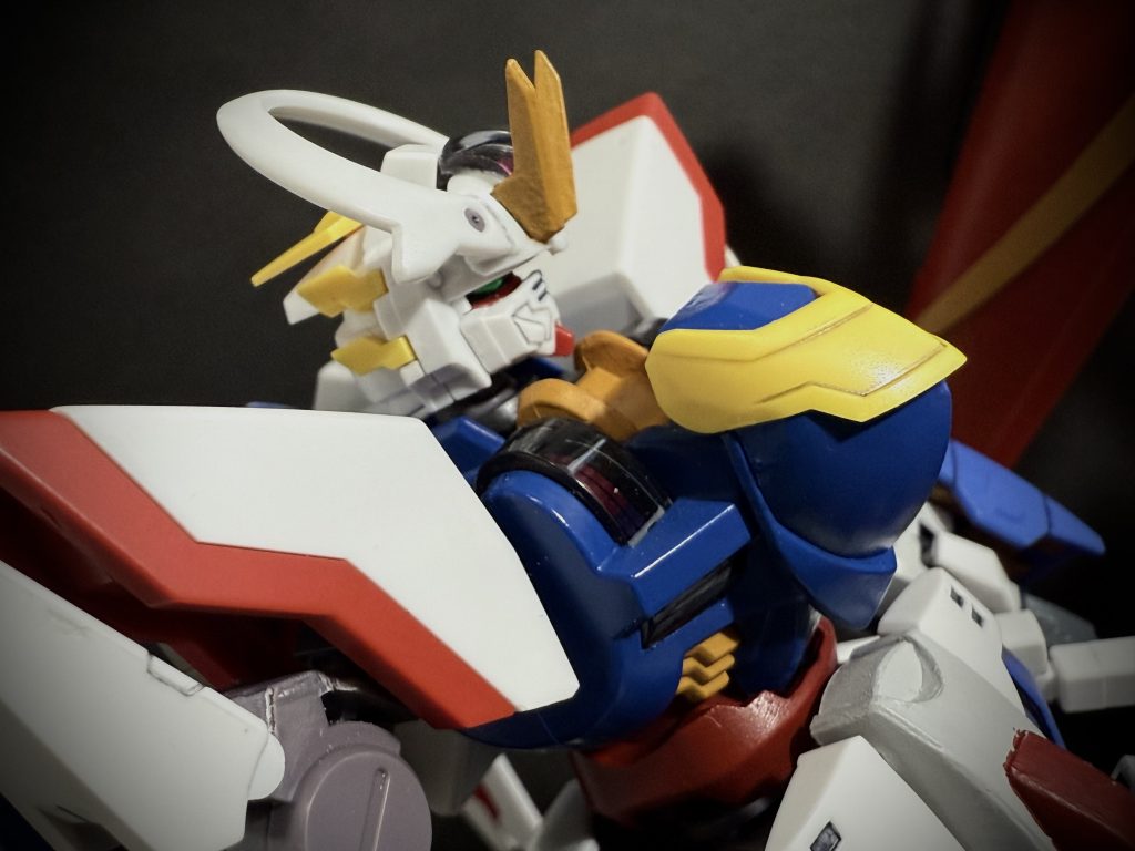 決闘　…後パイロット　バルゴ•ガルスキーMS &nbsp; ディランボルト（改修後）　穏やかな陽の光が樹々の隙間を縫って、ヤカンから注がれる液体。お湯をキラキラと輝かせている。十二時を少し回った今、林の向こう、アスティカシア校舎の方からは、賑やかな声が聞こえてくる。…いわゆる昼休みだ。慎重にカップ麺の中にお湯を注ぐ土門の耳に、何者かが近付いて来る音が聞こえてくる。…今、土門は学園敷地内の林の中でキャンプをしている。転校初日に決闘を行うという、前代未聞の事をやってのけた彼だが、彼を受け入れる地球寮としては、『人』を受け入れるより先にMS。シャイニングガンダムの受け入れに翻弄するハメになったのだ。…その結果、決闘後の機体の修理、整備と忙しく、肝心の『人』、土門に対し、まだ部屋の準備が出来ていない。という。つまる所、学園を通して見ても地球寮は人が足りないという事なのだ。暫く、何処かで寝泊りしてくれと言う地球寮の生徒の言葉に、修行で世界を回ってきた時の荷物のまま学園にきた土門は二つ返事で了承、こうして、林の中にひらけた所を見つけた土門は、決闘が終わってからの数日、ここでキャンプ生活なのである。「…俺に、何か用か？」　カップ麺にお湯を注ぎ終えた土門が、背後から近付く気配に、後ろも見ずに言う。「…ふ、気付いていたか」　そう言って、木々の間から下生えを踏み締め、巨体が姿を現す。バルゴ•ガルスキー。先日の決闘の相手。不良グループのリーダーだ。大きなボール箱を抱えたバルゴは、のそりと土門の前に回り込むと、持っていた箱を下ろし土門のまえに座る。訝しめな視線を向ける土門だったが、「…その箱は？」　土門の問いに、バルゴは少し目をそらし言う。「…カップ麺だ。…その、テント暮らしをしていると聞いてな」　少し、照れくさそうに頬を掻くバルゴだったが、土門の顔を見据えると、続けて言う。「…謝罪をしにきた。…決闘のまえに、酷いことを言ってしまった」　そう、静かに言い頭を下げるバルゴ。土門はそんなバルゴの顔を一瞥すると、割り箸をぱきりと割り、カップ麺をかき混ぜながら言う。林の中に、周りの木々の合間に聞こえるように、「…もう、気付いている。お前たち、出て来たらどうだ」　はっ、と頭を上げ、辺りを見回すバルゴ。すると、木々の合間から、次々と不良グループのメンバーが出て来るではないか、どこか照れくさそうに頭を掻く者、土門とバルゴに軽く頭を下げる者、サブリーダーまでもがモジモジしながら出て来る始末だ。「…お、お前ら。付いて来たのか」　後を付けて来たのだろう。バルゴは気付いていなかったらしい。狼狽するバルゴの様子をどこか微笑ましいと思いながら、土門はカップ麺を啜り言う。「…なぁに。気にしちゃいないさ。…それより」　土門はバルゴの脇に置かれた箱を指差すと、「…まったく、俺たち学生には、大事な昼メシの時間だろ。雁首揃えて俺の所に来なくてもいいだろうに。…どうだ、良ければ、みんなで食っていかないか？」　そう言って、傍のヤカンを掲げるのだった。〜数週後、地球寮　夕刻、寮の前で鍛錬をする土門に声をかける者がいる。「お前ら…」　長く伸びた影に土門が気付き顔を上げると、そこにはバルゴと、グループのサブリーダー、レスターシャが立っている。「あら、二人とも…、あ、もしかして、完成したの？」　土門が振り返ると、玲院が小走りでこちらに走ってくる。そのまま、レスターシャから持っていたタブレット端末を受け取ると二人、画面を覗き込みながら、何かを話し始める。そんな二人の様子を横目に土門はバルゴの横に行き。「仕上がったのか」「…ああ、細かい調整をお前達に手伝って欲しくてな」　そう言うバルゴの声が聞こえたのだろう。「全く、何処かの誰かさんが、近接戦闘に目覚めてしまってな…」　呆れたようにレスターシャが言う。玲院から手渡された端末を覗き込むと、画面には改良されたディランザが映し出されている。派手な装飾が取り外され、機体色はシックな黒を基調としたものに、両肩の大型シールドは、球体が埋め込まれた様なデザインの小型のものに変更されているのが目立つ。…機体名、『ディランボルト』。「火器の類は取り払った。…これがメインの武装だ」　そう言って、レスターシャの細い指が画面をスクロールさせると、小型化された肩のシールドに埋め込まれるように搭載された球体を指差す。隣に来たバルゴがレスターシャの話をつなぐ、「グラビティハンマーという。…これを振り回す、これで複数のレンジに対応できる」　これを振り回すために機体のセッティングを手伝って欲しい。そう言うバルゴを見上げながら土門は言う。「ああ、任せろ。…完成した時は」「…決闘（ファイト）だな」　続けて、バルゴは言う。「今度は、一対一でだ」「ああ！…望むところだ」　そう言って、バルゴと二人、拳を突き合わせて笑う土門。　〜少し離れた所で、夕陽の中、拳を合わせる男二人を、鼻息荒く見つめるレスターシャに気付いた者は、誰もいない。　　