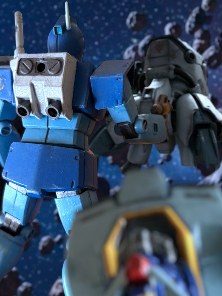 　正面のモニターは、ノイズで欠けているものの、まだ生きている。敵も、最後の武器の自壊で残っていた左腕が吹き飛んいる。もはやダルマ同然だ。いや、もう、頭もない。ただの鉄塊だ。青いジムが、鉄塊に素早く向かい、コクピットあたりにゴリっと、ビームスプレーガンの銃口を押しつけた。「ローワン、待て。」殺すなよ、と、通信を送る。『大尉、ご無事で。』「無事なものかよ、これが。」　敗けた。完敗だ。　経済力を由来にした機体性能の差。それを、見事に覆して、致命打を与えられ、今や自分は行動不能だ。　マイセンは、ノーマルスーツ用のバーニアを手早く装着し、ハッチを開け、機体の外に出た。ローワンが、危険です、と喚くのが通信機に聞こえた。「お前が守れ。なら、大丈夫だろう。」　フン、と、マイセンは穏やかに笑った。「パイロットは生きていると思う。コクピットを開けてやれ、敵ながら天晴れな、相手の顔を見てやろう。」　良い戦いをした。チタン系合金の精製技術を持たないジオンの機体で、コクピットブロックが残っているというのだから、敵のパイロットはなかなかに武運のあるヤツらしい。そして、ガンダムを打ち倒した。まるで、戦場に愛されて生まれてきたヤツのようではないか。戦士としての誇りを持ち続けてきたマイセンにとって、どんなヤツなのか興味を禁じ得ない相手だった。「小銃は持っている。大丈夫だ。拘束は、する。」　了解、とローワンは応える。『８２５、８２６、大尉がデブリ片に当たらないように、障壁になれ。』　付近の味方に指示を送るローワンの声が、スピーカーから聞こえた。次いで、ローワンのジムのマニピュレーターが、ダルマになった敵機の装甲を引き剝がすと、敵機のコクピット内の空気が、勢いよく吹き出す。噴射し切ったのを確認すると、マイセンは注意深く小銃を構えながら、剝き出しになったコクピットを覗く。中では、若いパイロットがぐったりとシートにもたれていたが、マイセンの姿を見ると、すぐにすっと姿勢を正した。濃い色のバイザー越しにも、何かを悟ったような、澄んだ瞳の色がよく分かった。　自らシートベルトを解き放ち、両手を上げながら、マイセンに近づく。マイセンは、投降の遺志表示と理解した。