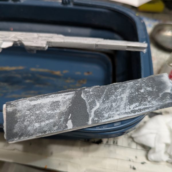 手を抜くのを推奨するのはプラモ制作としてどうなのでしょうか?手を抜くという表現は止めます。自分なりに愛情は注いでいますので・・・。良い例えがありました。料理愛好家の平野レミさんをご存知ですか?私もあの方の料理へのアプローチに準え、ガンプラ愛好家という事にします。モデラーっていうとなんか敷居上がっちゃいますからねwさて続きです。【ランナーからの切り離し】 とりあえずランナー塗装を済ませました。たまに思うのですが、パーツの裏面の色ってドムなら赤だと思いますが、他のMSって大体黒に塗ることが多くないですか?車のボンネットの裏も大体ボディと同じ色じゃないですかね?なので今回は裏面は塗らないです。では、ランナーから切り離しする前には何もしないかというと作業はします。モールドの掘り直しです。使う道具は100均のです。画像の赤い部分はキットに元からあるモールドです。青い方は特にモールドはないですが、掘っておくと墨入れが映えます。赤い方だけでも効果があります。切り離す時はプラモ用の鋏でざっくり切って、精密度の高いニッパーで余分を切るニ段階で対応します。仕上げ用のニッパーは私の持っている工具の方では高めです。ゴッドハンドの片刄ニッパーは壊しそうなので、タミヤのを使っています。3,000円〜4,000円くらいのです。正直高いかと思いますが、ゲート跡を綺麗に処理できるので、作業効率を格段に上げてくれる超便利アイテムなので、ここは頑張って買いましょう。塗装派も無塗装派も同じです。ちなみに私のタミヤニッパーは2代目です。初代は15年以上頑張ってくれました。中のバネが切れました。バネを入れれば復活するかもしれませんが、引退させて道具箱の中で眠っています。【合わせ目消し】ガンプラの大体の宿命として武器の合わせ目ですかね。基本モナカ構造なので、どうしても合わせ目が生じます。特に今回のように長めのライフルは特に合わせ目が発生しやすいです。ここはランナーを見た時から覚悟していました。(合わせ目消しのやり方)1.両面に接着剤(タミヤの白瓶)をドバッと塗り、塗ったあと1〜2分くらい待って「おりゃー」の掛け声と共にくっ付け、接着剤をムニュッとはみ出るまで圧着し、十分に乾燥(1週間なんて言いますね)させてからヤスリで削る。このやり方が一般的ですが、時間がかかるので今回は不採用です。2.合わせ目をそのままにする.変な話、合わせ目上等であれば無視していいかと思います。架空兵器なので、我々がおかしいと思っている合わせ目ですが、実は普通のことなのかもしれないからです。だからどうした!と思えば作業時間は大幅に短縮できます。3.段落ちモールドにする.ここで画像を紹介できませんが、筋彫り堂から出ている段モという工具を使うと段落ちモールド化させて分割線(合わせ目)を有効活用する。4.プラ板とかプラ棒を使って誤魔化す。プラ板を貼ったりして合わせ目を隠しちゃいます。私は最近は3と4の手段を使います。ということで画像はないですが、武器を早めに作っておく事にしました。説明書では武器は比較的最後に作ることが多いですが、最後に合わせ目消しが来ると萎えるので、私は複数を一気に作る時などは先に武器から作ったりします。ちなみにヤスリは水研ぎにしています。削りカスがヤスルと結構出ます。塗装の時よりも気を使ったほうがいいと思います。私はガスマスクみたいなのをつけています。間違って付けたままで宅配便を受け取りに行ったらびびられましたwその3へ続くー。(キャリバーンはバックパック以外は組めています) (6枚目)