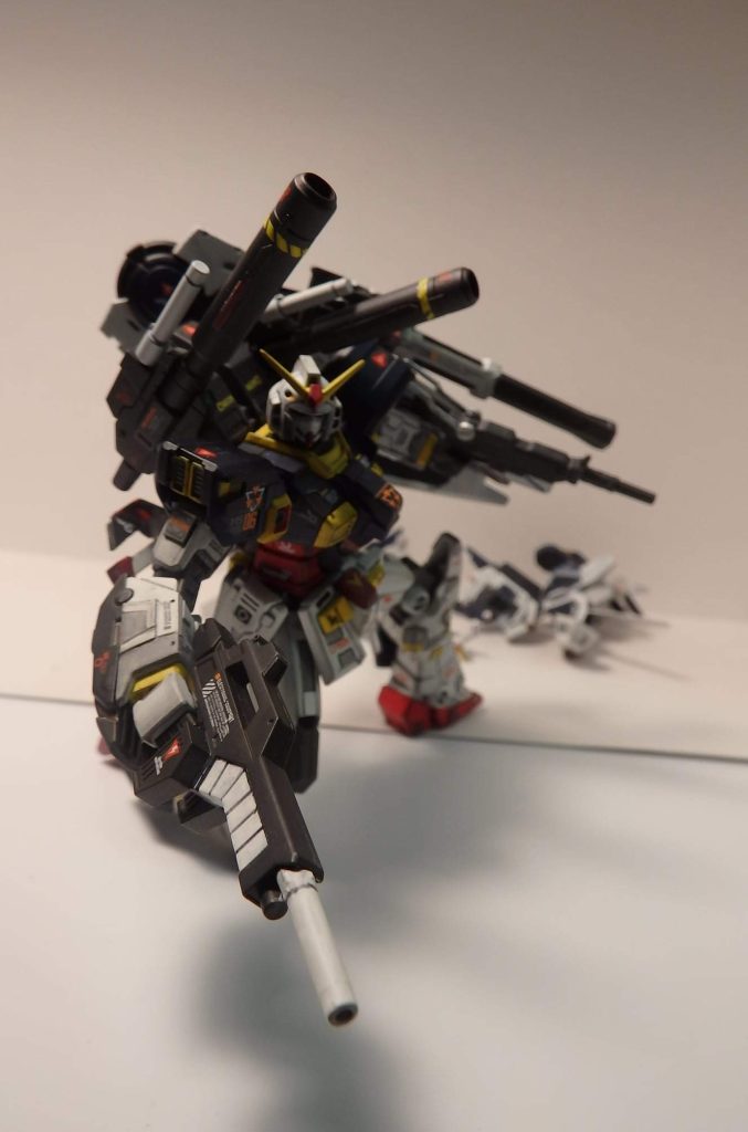 解説:東京湾付近のジャンク広場に埋もれていたモビルスーツのフレームを付近の修理工場の若社長が2万と言う破格な値段で購入、それを今まで集めてきたジャンクパーツをくっ付け、それを自称「ガンダム」と読んだ。ところが、機体は異世界で使われた魔法道具「巨神鎧(きょしんがい）」の成れの果てであり、それを求める異世界人や、それを本当にガンダムではないかと狙う軍事組織や者たちを掻い潜り、ようやくこの形になった。現在の姿は辛うじてスポンサーになってくれた三ッ橋重工業が製作した試作パーツを装着した姿である。