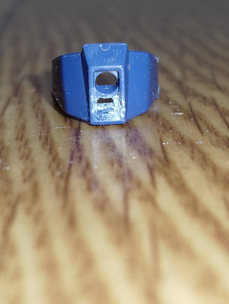 　精密狙撃用バイザーの加工です。　コチラのパーツ、上部の丸いセンサーはクリアパーツで再現されていますが、下部の四角い小さなセンサーはシールで補うようになっています。　しかしながら、この精密狙撃用バイザーはジム・スナイパーⅡのアイデンティティ。　なんとかリアルに仕上げたいと考え、下部の四角いセンサーをピンバイスとデザインナイフで切除し、裏から小さく切り出したクリアーグリーンのパーツを差し込んで再現することにしました。　というワケで、まずはご覧の通り、バイザーに四角い穴を開けます（細部はパテで微調整しました）。