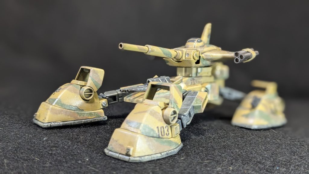 死の大地サハラ砂漠。その砂漠の中にある鉱石発掘基地に1発の砲撃音が鳴り響く。 175mm無反動砲を改良、冷却機能を付けた砂漠使用だ。其処から発射される成型炸薬弾(HEAT)が奥の指令塔を貫いた。 流石に2発も撃てば、いかな怠惰な連邦兵士でも砂漠の丘陵の影からの砲撃に気が付いた。 4本のアームを下げて高位置からの射撃姿勢を解除すると、周囲で爆発が起こり砂の柱が乱立した。 4本のアームの先に装備されたホバーユニットが鳴動し、その鉄の蜥蜴が砂塵を巻き上げ飛び出した。 甲高いターボファンの音が周囲を恐怖で包んでいく。 マゼラ・ホバー。ジオン公国の駆逐戦車マゼラ・アタックを砂漠用に魔改造された砂の戦場を駆けるバジリスク。 斜め前方にスライド走行をしながらマゼラトップでは回転しなかったターレットを回転させ、マゼラトップの翼端に有るロケットランチャーから全弾6発のロケットが無誘導で飛んでいく。 それらは61式戦車の出て来た格納庫に吸い込まれ、戦車中隊を格納庫共焼き尽くす。