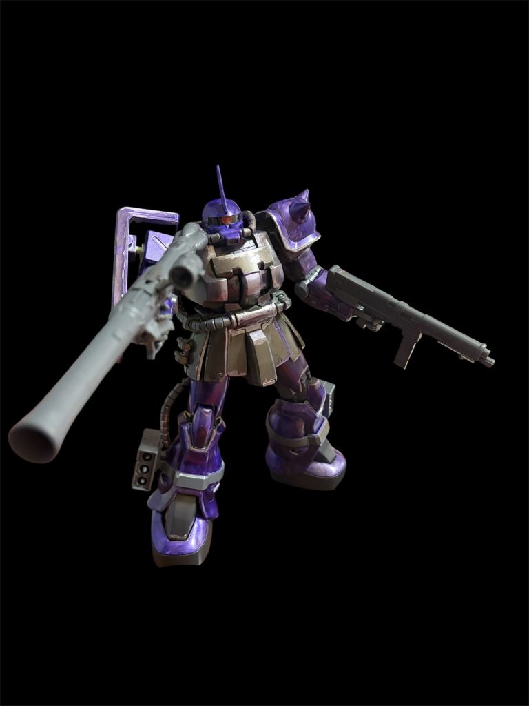 2. 質感とポージング• 金属的な重厚さ: 胸部や関節、武器（バズーカ、マシンガン）には、マットなメタリックグレーやチタン系を採用。外装のメタリックバイオレットとの質感のコントラストを強調しています。• 威圧的な立ち姿: ザク・バズーカとMMP-80マシンガンを両手に装備した重武装ポーズ。F2型のマッシブなシルエットを活かし、戦場で敵を圧倒する「エースパイロット機」の風格を表現しています。&nbsp;
