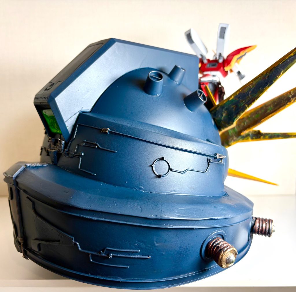 下半身のガンダムヘッドは、ジャンクパーツをあれこれ接着しました。見てわかる通り、電球のプラグ部分をおゆまる君で型取りしたヤツを接着しました。その根元の丸いリング状のパーツは、ガンプラなんかのランナー中央部の部分を切り出し、薄い輪っかになる様にヤスリで削り出ししたものを接着しました。