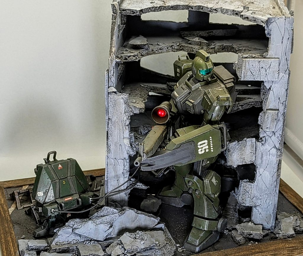 作品の設定は、廃墟のビルの陰に隠れて、ターゲットを狙撃するタイミングを覗っている場面です。スナイパーが狙撃しているその瞬間はもちろんカッコイイのですが、狙撃ポイントで待機しているところも自分は見せ場だと思っています。パイロットが抱えている、一発勝負の緊張感が伝わればいいなと思います。ジムスナイパーの大型狙撃ライフルは外部からのエネルギー供給を前提にしているそうなので、家に転がっていたジャンクパーツをかき集めてジェネレーターを自作しました。HGのEz8に付いているパラシュートパックをメインに作ったのですが、想像以上に見た方から正体を言い当てられるので驚きました（笑）モデラーの皆さんの知識と経験には脱帽です。
