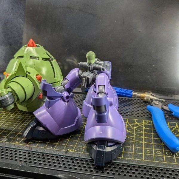 一番緊張する「異規格キット同士の接続軸改造(ミキシング)」に着手しました。今回は、リック・ドム(GQ)の腰部とゾックの上半身を接続するため、双方の元の接続ピンを思い切って切り落とし、新たにピンバイスで1mmの穴を開けて真鍮線を芯として通しています。この工程において一番苦労したのは、「とにかく穴位置をズレずに貫通させること」でした。相手は巨大なゾックのパーツ。1mmという極細の線とはいえ、コンマ数ミリでもピンバイスの角度が狂えば、パーツ同士に不自然な隙間ができてしまうため、文字通り息を止めるような思いで慎重に穴を開けました。結果的にズレることもなく、無事に真鍮線を通すことに成功!固定にはGSIクレオスの「Mr.ジャスト(はけ塗り瞬着)」を使用しています。このような繊細な軸打ち箇所において、はけ塗りタイプは液が余分なところに垂れたり白化したりする心配がなく、真鍮線の周囲の隙間だけにピンポイントで流し込めるため非常に重宝しました。真鍮線を芯にして強固に接着したため、強度も申し分なくガッチリと留められており、この仕上がりには大満足しています!(1枚目)
