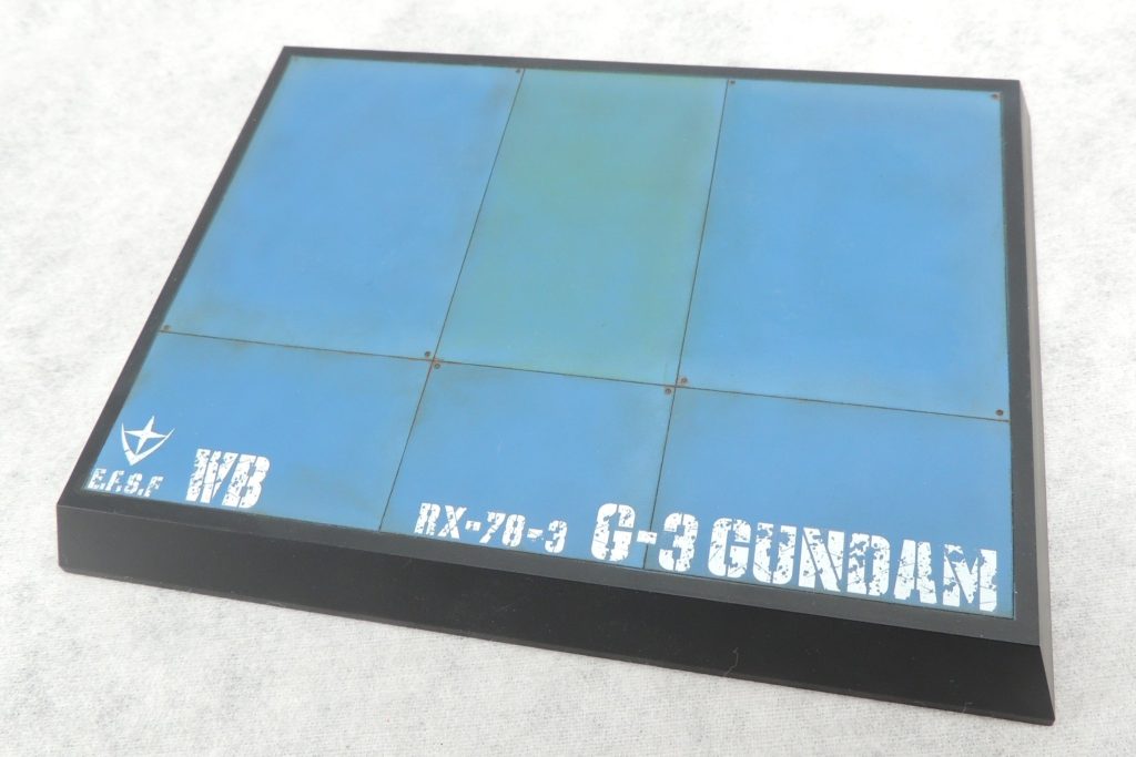 ■台座　台座はタミヤのディスプレイベース・スモール(148×108mm)を使用しました。スジ彫りやピンバイスによるネジ穴といったディティールを加えた後、枠をフラットブラック、天面を東雲ブルーで塗装し、Mrウェザリングカラーフィルタリキッドのスポットイエローやシェードブルーを薄く塗って色に深みをもたせてみました。　デカールはスジボリ堂のロボデカールからチッピングアルファベットとチッピングナンバーを使用しています。地球連邦軍のマークはガンダムデカールからチョイスし、乾燥後、チッピングデカールに合わせてデザインナイフで傷を入れました。
