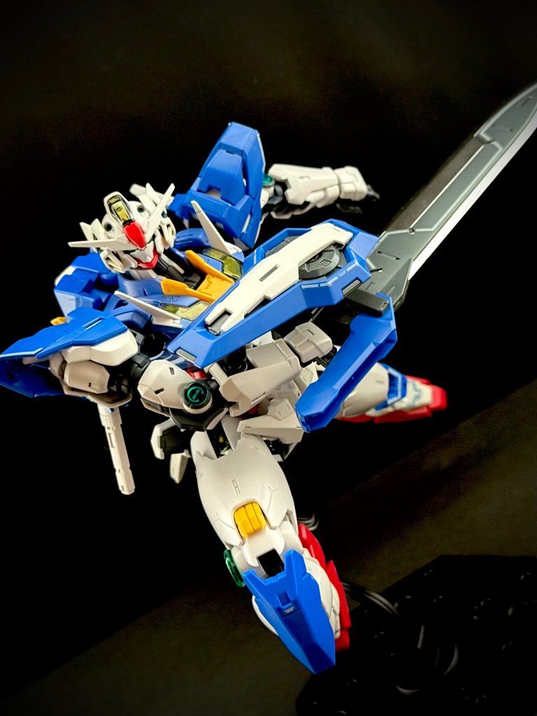 「…スレナ•M•セイエイ、目標を確認。…ガンダムエクシアル、これより争いを、根絶する」　それは、突然現れた。　なんでもない、いつもと変わらない決闘。くだらない生徒同士の諍いから始まった、いつも通りの決闘の最中だった。…「それ」は、現れた。　学園に設置されたスクリーンを、自身の端末を、そして直接その決闘を見ている全ての者の前にそれは現れた。　睨み合う、ザウォートとディランザの間に、「それ」、そのMSはふわりと着地すると、その右腕に折り畳まれていた巨大なブレードを展開、二機のMSに突きつけると、言う。「…任務了解。…これより、目標を駆逐する」　困惑するザウォートとディランザのパイロットをよそに、謎のMSは、姿勢を低くし、滑る様にディランザの方へ突進する。それと同時に、全ての生徒の端末に、全ての学園のスクリーン、モニターに、一瞬のノイズの後、一人の男。一人の奇妙な仮面を被った老人の姿が映し出される。学園内にいた者にはわからなかったが、この時、全ての地球圏内のあらゆるメディア、モニターというモニターに同じものが映し出されている。強力な電波ジャック。　全てのモニターの中でその老人は言う。「地球で、この宇宙で生まれ育った全ての人類に報告させていただきます。私はプロスペラ•シュヘンベルグ。…私達は、ソレスタアースビーイング」　何が起きているのかわからないディランザのパイロットは、動けない。一瞬で間合いを詰めた謎のMSは、すれ違いざま、ディランザの片手片足を、まるでバターを切るかの様に切り落とす。…モニターの中で老人は話続ける。「…機動兵器GUND-ARM （ガンダム）を所有する私設武装組織です。…私達、ソレスタアースビーイングの活動目的は、この世界から戦争行為を根絶する事にあります…」　ディランザの片手片足を切り裂いたガンダムは、そのまま背後に回り込むと、ディランザの残ったもう片方の手足も切り飛ばす。両足を失ったディランザ。そのまま地面へと倒れ込む。…？いや、倒れない。…“ガンダムの前にいる両手足のない胴体だけのディランザが浮いている“のだ。その様子を、正面から“見て“いたザウォートのパイロット。彼は気付く。ディランザが浮いているのでは無い。“ガンダムが片手で重量級のディランザの襟首を持ち、掴み上げているのだ“。…なんという出力（パワー）。唖然とするザウォートのパイロット。彼の耳には、ソレスタアースビーイングを名乗る老人の声しか聞こえない。「…只今をもって、全ての人類に宣言します。…どの様な理由があろうとも、私達は全ての戦争行為に対して、武力による介入を開始します」　ガンダムは、両手足を失い、戦闘能力を失ったディランザを、“優しく、いたわる様に“地面に横たえると、ゆっくりとザウォートへ向きなおる。「…戦争を幇助する、国、組織、企業なども、我々の武力介入の対象になりなす。…私達は、ソレスタアースビーイング。この世界から戦争を根絶させる為に創設された武装組織です…」　ザウォートのパイロットは見た。一瞬のうちに間合いを詰められ、一閃したブレードにより、自分。自機の手足が切り飛ばされるのを。