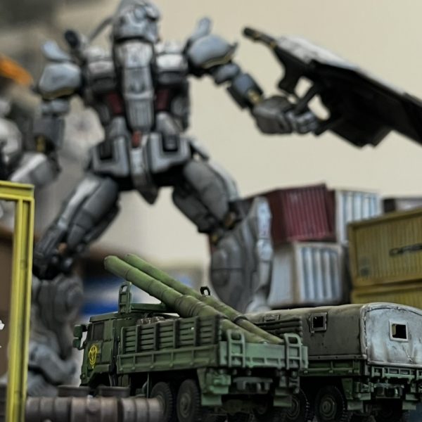 サムネのガンダムの背後からコンテナ越しにイリア・ソラリのザクがヒートホークで飛び掛かる瞬間を捉えたイメージでおります。ジオラマのレイアウトを調整しながら追加で必要と思われる小物をせっせと作ってます。ベースのサイズに合わせてちょっとした作業小屋を追加。その脇にトラック2台。その内の1台はオープントップにして荷台にマゼラの砲身を2本載せてます。（劇中でもそんなシーンがありました）自衛隊のトラックにジオンマーク！似合います😅ガンダムのシールドにはリューターでダメージ加工を施してザクマシンガンの弾痕をば表現。廃車も良い感じですがブレーキランプ等はしっかり塗装した方が良さそうです。コンテナは白の下の赤にも衝撃が伝わり下が少し浮き上がった状態にしています。その隙間からホコリが出ている表現を真綿で出来たら面白いと思っています。（3枚目）