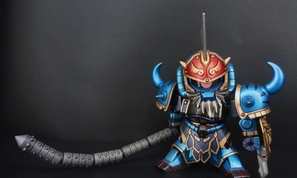 【作中での概要】​黄蓋グフ（こうがいグフ）は、『SDガンダム 三国伝』シリーズに登場する武将。孫権ガンダムらに仕える「長沙四天王」の一人。演者はMS-07B グフ。​孫堅ゼフィランサスの代から孫家に尽くす大ベテランの老将。かつては「長沙の赤陣」と呼ばれた一騎当千の猛者であり、老齢となった作中でも自慢の怪力と武勇は衰えていない。​劇中では、先代・孫堅の遺志を受け継ぎ、孫策サイサリスや孫権らを力強く支える後見人として活躍。巨大な鉄球型武器「多節鞭（鉄鎖流星）」を豪快に振り回す重厚な戦術を得意とし、命を賭して孫家の若武者たちを導く「東呉の頼れる鉄壁」として描かれている。※要約Gemini